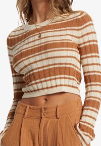 Brązowy i kremowy prążkowany sweter cropped o rypsowej fakturze. W zestawie z jasnobrązowymi spodniami z wysokim stanem, z podwójnym zapięciem na guziki.