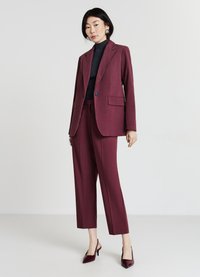 Completo bordeaux su misura con giacca attillata e pantaloni a gamba dritta. Il tessuto ha una consistenza liscia. Abbinato a scarpe con tacco scuro.