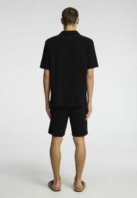 Czarna koszula z krótkim rękawem i dopasowane szorty wykonane z teksturowanej tkaniny, o luźnym kroju i płaskim kołnierzyku. Widoczne sandały.