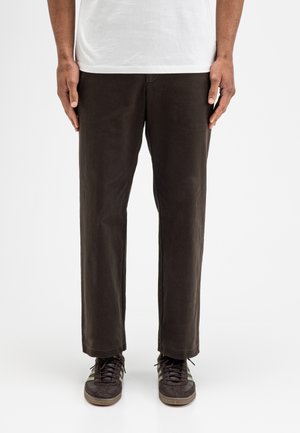 Person trägt dunkelbraune gerade geschnittene Hose, schwarze und graue Sneakers und ein weißes Hemd und steht mit den Händen an den Seiten.
