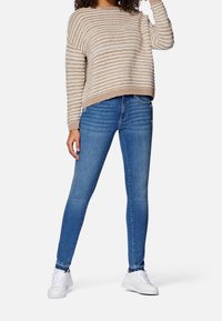 Beżowo-biały sweter w paski, luźny krój, zestawiony z dopasowanymi niebieskimi jeansami i białymi trampkami.