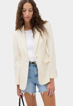 Kvinde med langt krøllet hår iført en cremefarvet blazer, hvid skjorte, slidte denimbukser og holder en sort taskestrop.