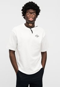 Λευκό βαμβακερό μπλουζάκι με κοντά μανίκια, με μαύρο λογότυπο "Jordan Café" στο στήθος, φορεμένο με σκούρο παντελόνι. Το μοντέλο έχει μαύρα μαλλιά με υφή.