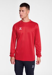 Maglietta sportiva rossa a maniche lunghe realizzata in tessuto morbido, con collo rotondo e un logo bianco sul petto, abbinata a pantaloni scuri.