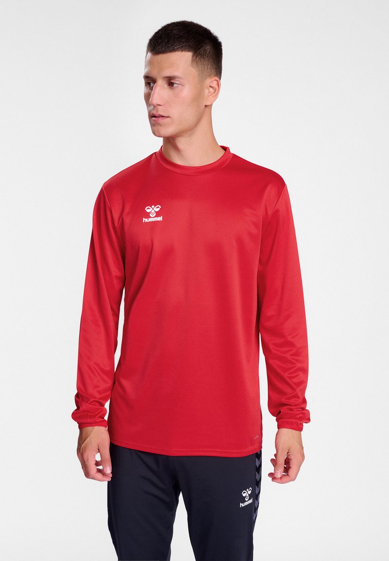 Maglietta sportiva rossa a maniche lunghe realizzata in tessuto morbido, con collo rotondo e un logo bianco sul petto, abbinata a pantaloni scuri.