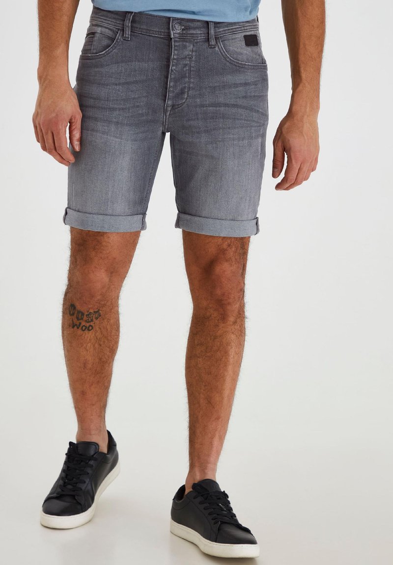 Mann trägt hochgekrempelte graue Denim-Shorts und schwarze Sneakers und zeigt ein Tattoo am linken Unterschenkel.