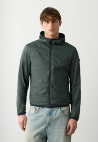 Dunkelgrüne, leichte Jacke mit Kapuze, Reißverschluss und elastischen Bündchen. Kombination mit hellblauen Denim-Jeans, neutralem Hintergrund.