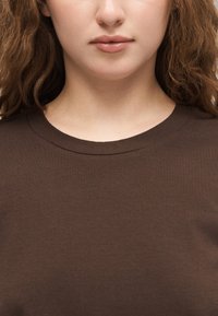 Persoană tânără cu ten deschis și păr creț, maro, purtând un tricou simplu maro închis cu guler rotund, buzele închise, expresie neutră.