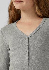 Close-up van een persoon die een grijze geribbelde V-hals shirt draagt met vier knopen aan de voorkant, waarbij de nek en een deel van het gezicht zichtbaar zijn.