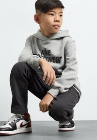 Pelēks kapucis džemperis ar melnu "Nike Sportswear" logotipu, apvienots ar tumšām biksēm un melniem, baltajiem un bordo sporta apaviem. Ceļgala pozā.