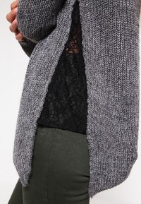 Persona che indossa un cardigan grigio a maglia sopra una maglietta nera in pizzo e pantaloni verde scuro, con le braccia incrociate dietro la schiena.