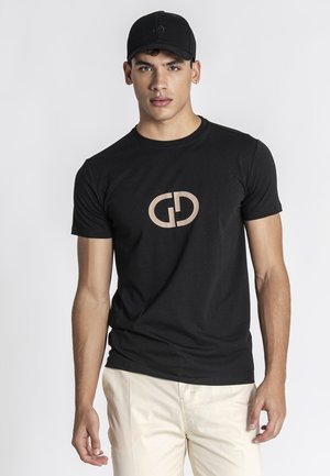 Mladić koji nosi crnu kapu, crnu majicu s bež logotipom i svijetlo bež hlače, stoji nasuprot jednobojnoj svijetlosivoj pozadini.