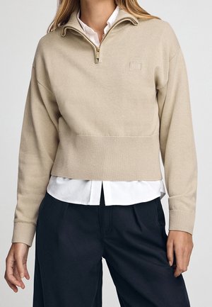 Femme portant un pull beige à demi-fermeture éclair sur une chemise blanche avec un pantalon bleu marine, debout devant un fond clair uni.