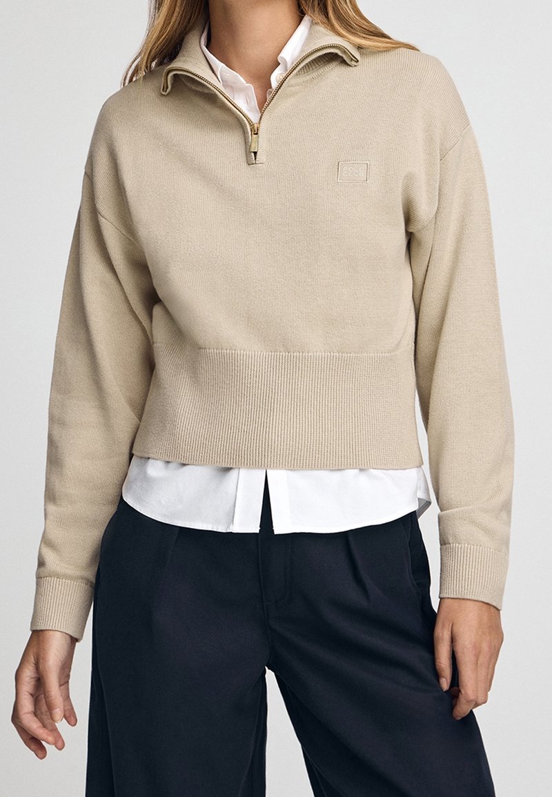 Femme portant un pull beige à demi-fermeture éclair sur une chemise blanche avec un pantalon bleu marine, debout devant un fond clair uni.