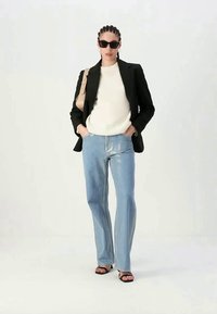 Mulher a usar um blazer preto, um suéter branco, jeans azul claro, sandálias pretas, óculos de sol e a carregar uma mala de ombro bege.
