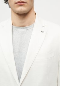 Gros plan sur une personne portant un blazer blanc par-dessus un t-shirt gris clair à col rond, avec une petite épingle argentée "MK" sur le revers du blazer.