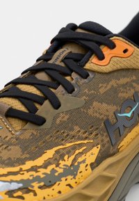 Scarpa da corsa marrone e beige con tomaia testurizzata, lacci neri e dettagli arancioni. Presenta un design decorato e una punta rinforzata.