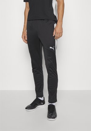 Osoba ubrana w czarne spodnie dresowe Puma z białymi paskami po bokach i czarne buty sportowe z białą podeszwą, stojąca na jednolitym tle.