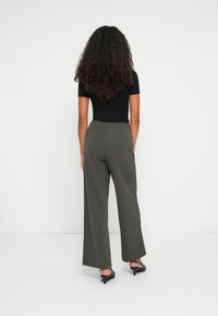 Eine Frau mit langen lockigen Haaren steht mit dem Rücken zur Kamera. Sie trägt ein schwarzes Kurzarmoberteil, weite olivgrüne Hosen und schwarze Absatzsandalen.