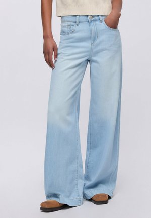 Persona che indossa jeans larghi azzurri con un top lavorato a maglia beige senza maniche e scarpe marroni, con una mano in tasca, su sfondo semplice.