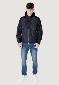 Giacca a vento blu navy con cappuccio, dotata di zip frontale e dettagli con logo. Abbinata a jeans azzurri e sneakers bianche.