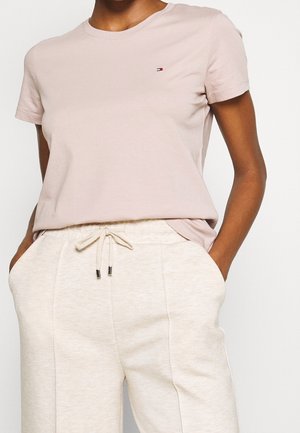 Person wearing a light pink short-sleeve shirt with small logo and cream-colored drawstring pants with hands in pockets.