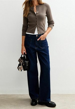 Žena nosící hnědý zapnutý kardigan přes bílý top, tmavé široké džíny, černé boty a držící malou černou kabelku s přívěsky.