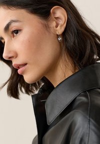 Silberner herzförmiger Ohrring, der vom Ohr hängt, getragen zu einer schwarzen Lederjacke mit weitem Kragen und glatter Textur.