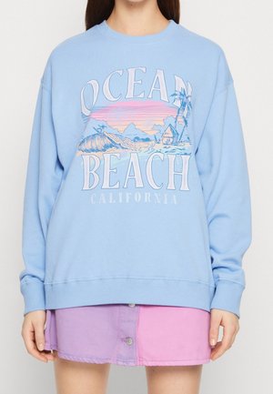 Osoba nosząca jasnoniebieską bluzę z napisem "Ocean Beach California" i motywem plaży, dopasowaną do dwukolorowej spódnicy różowo-fioletowej.