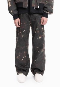 Person som har på seg løstsittende mørke kamuflasjebukser med lastebukinspirert design og hvite sko, med hendene avslappet ned langs sidene, stående foran en hvit bakgrunn.