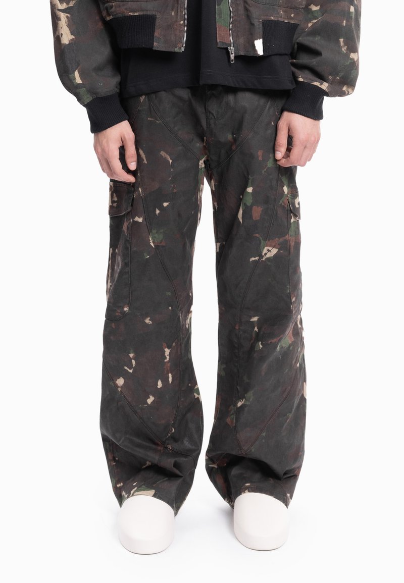 Person som har på seg løstsittende mørke kamuflasjebukser med lastebukinspirert design og hvite sko, med hendene avslappet ned langs sidene, stående foran en hvit bakgrunn.