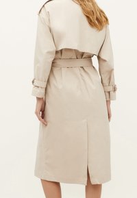 Beżowy płaszcz trencz z paskiem w talii, długimi rękawami, rozcięciem z tyłu i skróconym tyłem, wykonany z gładkiej tkaniny. Styl minimalistyczny.