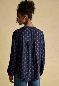 Tumši zila ziedu blūze ar garām piedurknēm, kurai ir mazs ziedu raksts rozā un zaļā krāsā, un plecu detaļa aizmugurē.