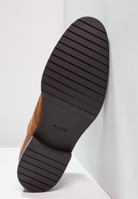 Semelle de chaussure en daim marron avec une semelle extérieure en caoutchouc noir texturé et des rainures proéminentes, arborant le nom de la marque "ALDO" embossé sur le côté.