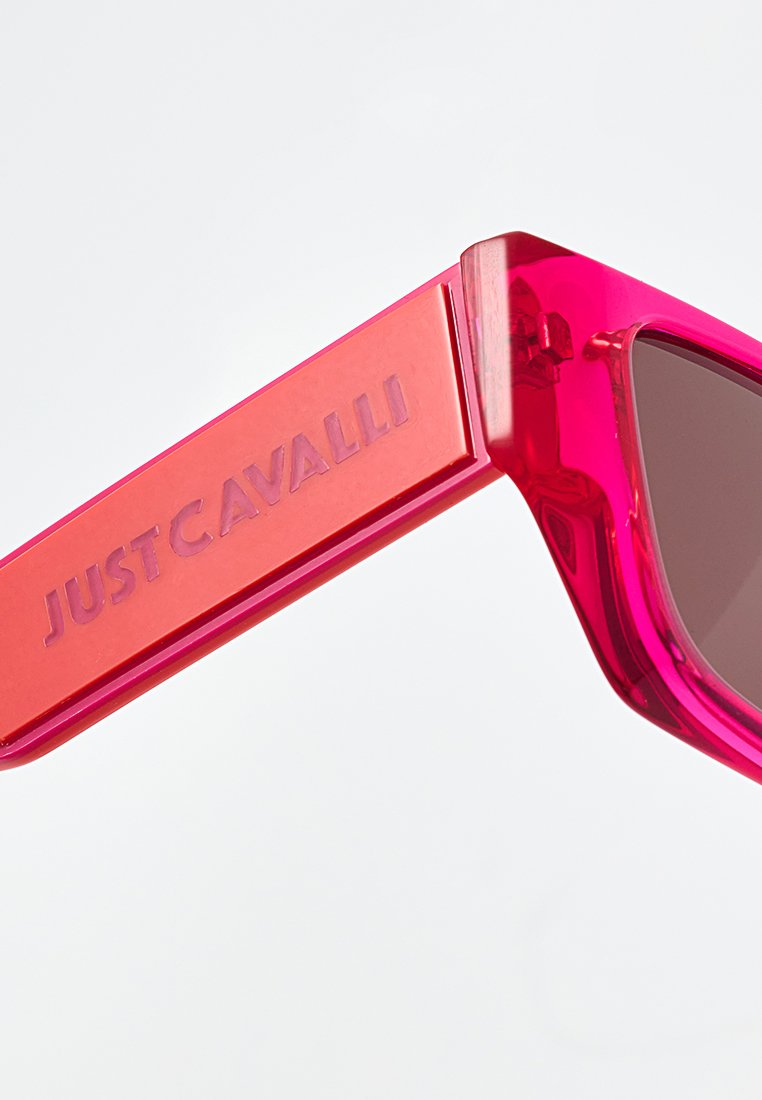 Lunettes de soleil transparentes roses avec des verres foncés. La monture présente un design audacieux et angulaire avec une large branche ornée d'une marque en relief indiquant "JUST CAVALLI."
