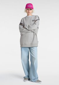 Grijze sweatshirt met "VANS" logo en strepen, gecombineerd met lichtblauwe wijde jeans en geruite slip-on schoenen. Roze pet met logo.