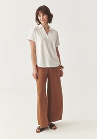 Witte korte mouwen polo met een V-hals, gecombineerd met losse bruine wijde broekspijpen en zwarte sandalen. Casual en ontspannen pasvorm.