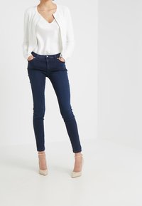 Biały kardigan z żebrowanym wzorem noszony na kremowej bluzce z dekoltem w serek, połączony z ciemnoniebieskimi jeansami typu skinny oraz cielistymi szpilkami z paskiem wokół kostki.