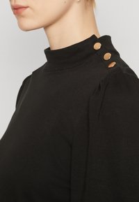 Black long-sleeve top with a high collar, featuring three decorative gold buttons at the shoulder and a slightly textured fabric.