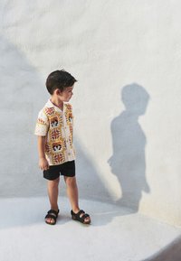 Kortärmad skjorta med en färgstark virkadesign i orange, gult och vitt, kombinerad med svarta shorts och sandaler.