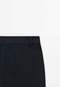 Gros plan de la ceinture arrière et d'une unique poche arrière horizontale d'un pantalon formel noir sur fond blanc.