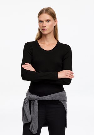 Femme aux cheveux blonds raides porte un haut noir à manches longues et un pull gris noué autour de la taille, debout, les bras croisés.