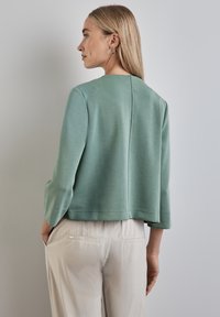 Sweatshirt cropped vert menthe avec des manches trois-quarts, texture lisse et détails de couture à l'arrière, associé à un pantalon de couleur claire.