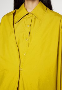 Femme portant une chemise jaune vif à boutons avec un grand col, montrée des épaules jusqu'au milieu de la poitrine sur fond blanc.