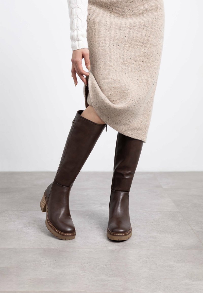 Botas de cuero marrón hasta la rodilla con un tacón grueso y suela de goma texturizada, combinadas con un vestido de punto beige y una manga de punto blanco.