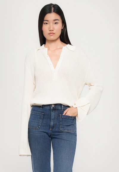 Pullover bianco a coste con colletto e patta dei bottoni, abbinato a jeans blu scuro con tasche anteriori e un design aderente.