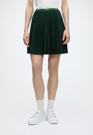Personne portant une jupe plissée vert foncé avec une ceinture à logo Lacoste, un haut blanc, des chaussettes blanches et des baskets blanches à plateforme, debout sur un sol blanc.