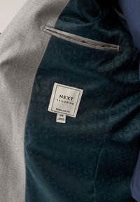 Doublure intérieure d'un blazer gris avec une poche sombre à motif et une étiquette « NEXT Tailoring Regular Fit 40R » cousue à l'intérieur.