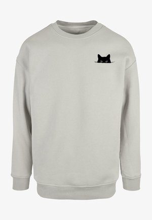 Sweatshirt gris clair en tissu doux, orné d'un petit graphique d'un chat noir aux grands yeux sur la poitrine gauche. Poignets et ourlet côtelés.