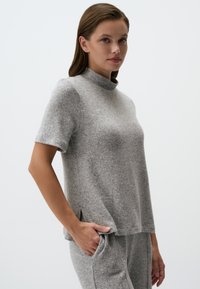 Grijze top met korte mouwen en een hoge kraag, gemaakt van zachte stof. Bevat zijsplitsingen en heeft een losse fit, gedragen met bijpassende grijze broek.